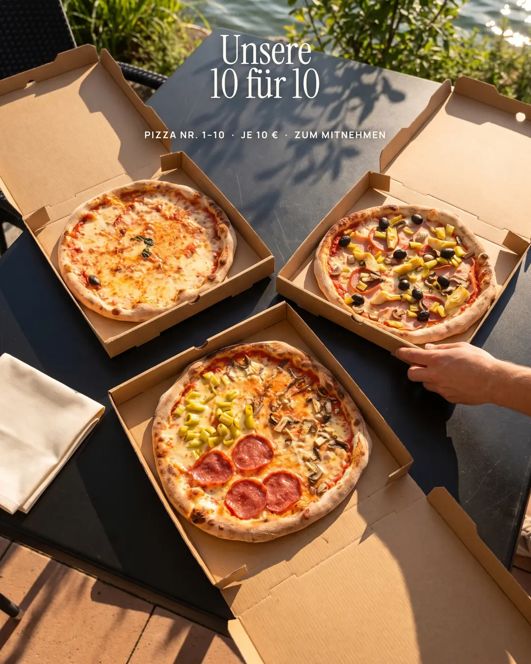 Drei Pizzen in geöffneten Kartons auf einem Tisch direkt am See.